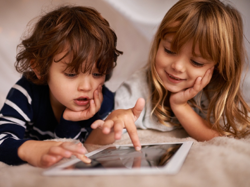 Lille mørkhåret dreng og lille lyshåret pige ligger og spiller på en Ipad.
