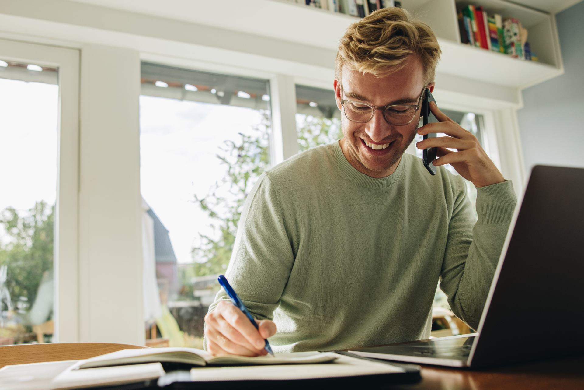 Ung mand sidder ved et bord med sin computer, mens han skriver ned i notesbog og taler i telefon.